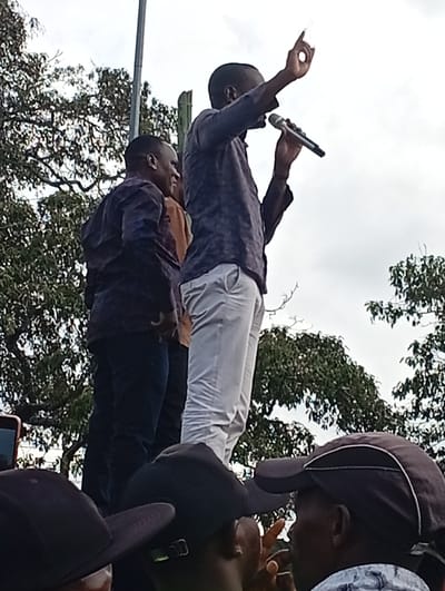 The image depicts an outdoor public gathering in Mukuyuni, Kenya. In the upper-center, a dark-skinned male speaker, dressed in a dark collared shirt and light-colored trousers, addresses an audience. He holds a black microphone in his left hand and extends his right hand upwards, forming an 'okay' gesture. To his left and slightly behind him, another dark-skinned male, wearing a dark patterned long-sleeved shirt and dark trousers, stands observing. A third individual in a brown shirt is partially visible between the two main figures. The foreground shows the heads of several individuals, some wearing dark baseball caps, looking upwards towards the speakers. A red-cased mobile phone is partially visible on the bottom left, held by a foreground subject. The background consists of green tree foliage and an overcast sky, with a tall, light-colored pole and a darker wooden pole erected behind the speakers.