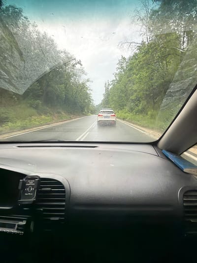 The image shows a view from inside a car, looking through the windshield. The windshield is wet, with raindrops visible. A white SUV is driving ahead on a wet, two-lane road lined with trees. The road appears to be in a rural or suburban area. The dashboard of the car is visible in the foreground, with a phone mount attached to a central air vent. The sky is overcast, suggesting rainy or cloudy weather. The location is identified as Bilka, Bulgaria.