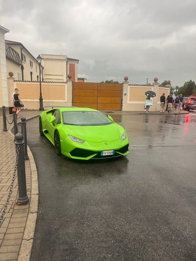 The image depicts a bright green Lamborghini Huracán parked on a wet asphalt road in front of a cream-colored building with a large wooden gate, likely in the city of Noventa di Piave, Italy, given the context. The sky is overcast and it appears to be raining, as evidenced by the wet surfaces and several people in the background holding umbrellas.
In the foreground, the Lamborghini is the primary subject, angled towards the right of the frame, showcasing its sleek design. Its license plate is visible and reads "FW-926WN". To the left of the car, a paved walkway with a chain-link fence and bollards runs alongside the road. A woman in a black outfit is walking on the walkway, carrying shopping bags.
In the background, beyond the wooden gate, a group of people are standing, some holding umbrellas. The building behind them has architectural elements such as decorative spheres on pillars and tiled roofs. Trees are visible in the far background. The overall scene suggests an outdoor event or a visit to a commercial area, possibly a designer outlet given the shopping bags, in a moderately urban environment under inclement weather.