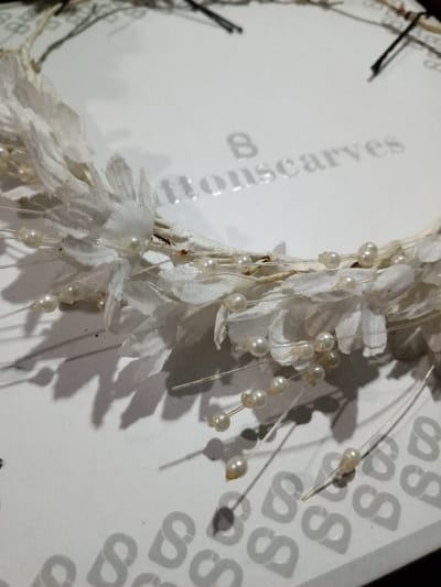 A close-up reveals a white, decorative wreath resting on a surface patterned with repeating "S" shapes. The wreath features delicate white fabric flowers interspersed with small, pearlescent beads attached to thin wires. Shadows cast by the wreath add depth, highlighting the texture of both the floral arrangement and the underlying surface. The inscription "Bittonscarves" is faintly visible in the background.