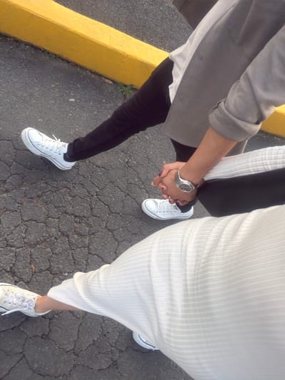An aerial perspective captures a couple holding hands while standing on cracked asphalt. The person on the left wears a gray jacket, white shirt, black pants, and white sneakers, sporting a wristwatch. The person on the right is dressed in a white ribbed dress and white sneakers. A yellow curb is visible in the image's left corner. The focus is on their connected hands and footwear.