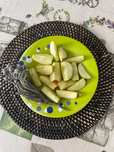 A top-down shot reveals a plate of sliced apples on a black woven placemat. The light green plate, speckled with blue and white circles, holds approximately fifteen apple slices, their pale flesh tinged with hints of green and red peel. A decorative gray tassel lies adjacent to the apples. The placemat sits atop a patterned tablecloth featuring illustrations of flowers, birds, and vases. The scene is likely located in the Лесной Пейзаж Кп (шарапово) area of Russia, based on the photo's origin.
