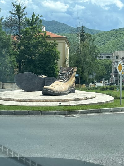 The image shows a large sculpture of two hiking boots located on a circular platform in the city of Peshtera, Bulgaria. One boot is upright and laced, while the other is lying on its side. The sculpture is situated in what appears to be a public space, possibly a roundabout or a park, with green grass and a paved road visible. In the background, there are trees, buildings, and rolling green hills under a cloudy sky. The overall scene suggests an outdoor public art installation in an urban environment surrounded by nature.