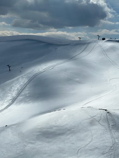 This image depicts a snow-covered mountainous landscape in Katun Bačko brdo, Montenegro, under a partially overcast sky. The scene is dominated by undulating terrain covered in white snow, marked with numerous tracks. Several ski lift poles are visible, primarily along the upper ridge of the mountains, indicating an operational or recently used ski area. Two distinct types of tracks are observable: narrower, winding tracks consistent with skis or snowboards, and wider, straighter tracks, possibly from snow groomers or snowmobiles, traversing the slopes. A small, dark structure, potentially a service building or shelter, is situated near one of the uppermost ski lift poles on the right side of the image. Sunlight intermittently illuminates sections of the snow, creating contrasting areas of bright white and cooler shadows. No individuals are visible. The sky features dark grey clouds interspersed with brighter patches.