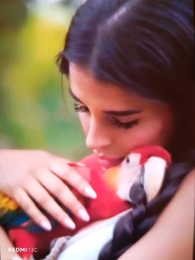 A woman with dark hair and long lashes embraces a vibrant parrot with red, yellow, and white plumage. Her fair-skinned hands gently hold the bird against her chest. The parrot is nestled closely, its beak near her face. The background is a blur of green and yellow foliage, suggesting a natural outdoor setting. A watermark "REDMI 13C" is visible in the lower-left corner. The image has a soft, slightly blurry quality.