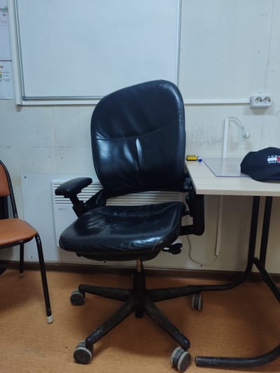 The image depicts an indoor scene, likely an office or study area, in Қосшағыл ауылдық округі, Kazakhstan. The central focus is a black leather office chair with a five-wheeled base, positioned in front of a light-colored desk. The chair appears worn. To the left of the office chair, a second chair with an orange seat and black metal frame is partially visible. The desk has a light wood-effect surface and black metal legs. On the desk, a clear plastic barrier, a black baseball cap with the Tommy Hilfiger logo, a blue pen, and a yellow eraser are present. In the background, a white whiteboard is mounted on the wall, and a white electrical outlet with two sockets is visible on the wall to the right, with a white conduit running along the wall. A white radiator is located behind the office chair. The floor is covered with a textured, tan-colored material. The lighting is artificial, contributing to a cool-toned ambiance.