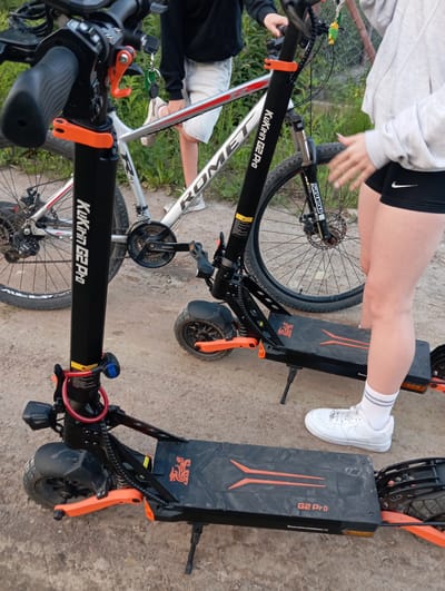 In an outdoor, dirt ground setting in Dybów-Kolonia, Poland, two individuals are visible near a bicycle and an electric scooter. The person on the right, wearing white socks and white sneakers, has their legs positioned over the electric scooter. They are also wearing black shorts with a Nike logo and a grey hooded sweatshirt. Their right hand is extended and placed on the seat of the bicycle. The person on the left is partially visible, wearing a black hoodie and white shorts. The electric scooter is black with orange accents and the brand name "KUKIRIN G2 PRO" is visible on its stem. A red lock is attached to the scooter's stem. The bicycle is silver with red accents, and the brand name "ROMET" is visible on its frame. The scene is lit by natural daylight.