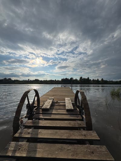 The image depicts a wooden pier extending into a calm body of water, likely a lake or river, in Sitno, Russia. The sky above is a mix of dramatic grey clouds with patches of bright sunlight breaking through, creating reflections on the water's surface. The pier, made of weathered wooden planks, has two rustic metal wheels attached to its sides near the entrance. These wheels appear to be part of a mechanism, possibly for launching or retrieving a boat. The far bank of the water is lined with a dense forest. The scene is serene and natural, with no people visible. The overall impression is one of a quiet, somewhat remote waterfront location.