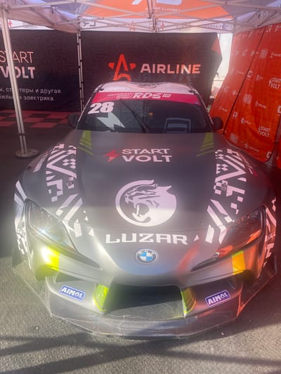 The image shows the front of a grey race car with various decals and sponsorships. The car is parked under a white tent structure, with banners visible in the background. The banners are predominantly orange and black and feature logos and text, including "START VOLT," "AIRLINE," and "CARVILLE."
On the hood of the car, there is a large decal with a stylized animal, possibly a panther or jaguar, and the text "START VOLT." There is also a number "28" on the windshield, along with other racing decals. The front bumper of the car has carbon fiber accents and two "AIMOL" stickers. The car has yellow and green accents on the headlights and front splitter. The ground beneath the car appears to be a textured dark surface, possibly a mat or flooring, with shadows cast from the tent structure.
No people are visible in the image. The environment suggests a race event or a car show. The location specified is the city of Берёзовка, Russia.