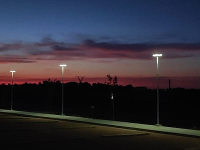 The image captures a twilight scene in the city of Valverde de Leganés, Spain, with a darkening sky displaying hues of deep blue transitioning to pink and orange near the horizon. Three modern streetlights with white LED fixtures stand tall, illuminating a paved area in the foreground, possibly a parking lot, marked with yellow lines. The landscape behind the streetlights is silhouetted in darkness, with a dense line of trees and vegetation forming the background. A faint structure resembling a power line pylon can be seen against the sky. The overall atmosphere is serene and dimly lit, characteristic of the early evening hours.