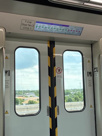 The image shows the interior of a train car, likely on the MRT Pink Line in Bangkok, Thailand, as indicated by the station information displayed on a screen above the doors. The screen shows "Next Station: Bang Rak Noi Tha It" and "Destination: Tao Poon," identifying the current location and upcoming stops. The train car features two doors with windows, through which a landscape of houses and greenery can be seen. Warning signs and door operation instructions are visible on the doors. One sign explicitly states "DO NOT LEAN AGAINST DOORS." The overall scene is clean and functional, typical of public transportation.