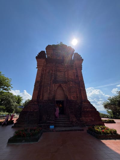A low-angle, full-frame shot captures a weathered, ancient brick tower against a bright blue sky. The sun is high and partially obscured by the top of the tower, casting a strong lens flare. The tower, constructed of reddish-brown bricks, has a stepped, pyramid-like base that narrows as it ascends. The top of the tower is uneven and broken, with some vegetation growing from its apex. A dark, arched entrance is visible at the base, with steps leading up to it. A person in a pink outfit stands on the steps to the right of the entrance. To the left of the tower, several people in traditional Vietnamese clothing are visible, walking on a paved area. The ground is paved with reddish-brown tiles, and planters filled with red flowers and green foliage are placed around the base of the tower. Lush green trees flank the scene on the left and right. The overall impression is one of historical significance and cultural exploration in a tropical setting. The location is Po Nagar Cham Towers in Tuy Hoa Ward, Vietnam.