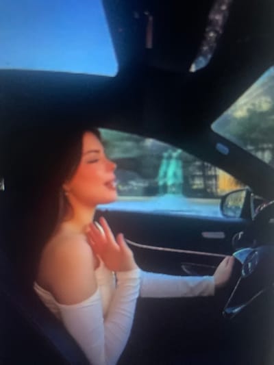 The image shows a young woman with long, dark hair sitting in the driver's seat of a car. She is wearing a white off-the-shoulder top with long sleeves. Her head is turned to the side, and her eyes are closed, with her mouth slightly open as if singing or speaking. Her right hand is raised, with fingers splayed, near her face. The car has a sunroof open, revealing a blue sky. The interior of the car is dark, with the steering wheel visible. Outside the car, through the windshield and side window, there are blurred green and yellow shapes that appear to be trees and buildings. The lighting suggests it is daytime. The image quality is poor, with significant blur and low resolution. The location in the city of Kołobrzeg, Poland, is not discernible from the visual information provided.