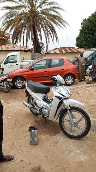 The image shows a street scene in Kawo, Nigeria. A silver INCHENG motorcycle is parked on a dirt patch with a plastic jerrycan nearby. Behind the motorcycle, there is an orange Peugeot 206, a white utility truck, and another motorcycle. Two men in traditional attire are standing near the Peugeot, with their backs turned toward the camera. The background includes buildings with corrugated metal roofs and a large palm tree. The sky appears overcast.