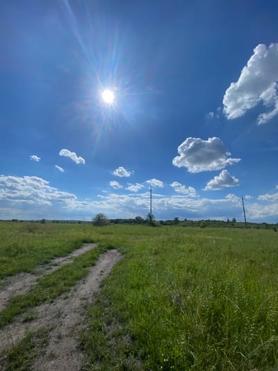 The image shows a dirt road leading through a grassy field under a bright blue sky with scattered white clouds. The sun is prominently visible in the upper left corner, casting a strong glare and lens flare. Power lines and utility poles are visible in the background, stretching across the horizon. The landscape appears to be rural, with some scattered trees and vegetation in the distance. The overall impression is one of a sunny, open, and somewhat undeveloped area.