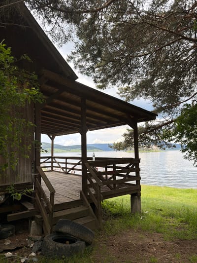 This image captures a rustic lakeside scene at Цигов чарк, Bulgaria. The primary subject is a weathered, dark-stained wooden building with a pitched roof, situated directly on the edge of a large body of water. A covered wooden porch extends from the building, featuring a plank floor, a wooden railing with criss-cross balustrades, and visible roof rafters. Wooden steps ascend to the porch from the immediate foreground. The structure is elevated on concrete blocks and timber supports.
In the foreground, the ground is a mix of bare earth and sparse grass. Two old, dark car tires, one lying flat and another propped against it, are discarded near the steps. Scattered debris, including wooden scraps, concrete fragments, and small pieces of trash, are visible beneath and around the base of the wooden structure. Lush green grass covers the ground closer to the water's edge.
The midground and background are dominated by a calm body of water, likely a lake or reservoir, reflecting the sky. Across the water, distant forested hills or mountains are visible. A white, indistinct vessel or structure is present on the water's surface in the middle distance. Further along the opposite shore, a small landmass or island with a cluster of white, possibly bare, trees or structures can be seen.
Overhead and framing the scene, dense branches and needles of a conifer tree, likely a pine, are prominent, partially obscuring the partly cloudy sky. Green leafy vegetation grows on the left side of the wooden building.
On the porch, a clear plastic water bottle rests on the railing on the right side. A white lifebuoy is hanging on the interior railing of the porch. Faint, indistinct human figures are discernible in the background, positioned behind the left section of the porch railing. The sky is partly cloudy with variations in brightness, suggesting an overcast or partly sunny day. No direct human actions or interactions are clearly depicted within the main frame.