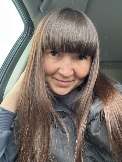 A woman with long, straight brown hair and bangs is seated in a vehicle. She is wearing a black jacket over a gray sweatshirt. The woman is smiling slightly at the camera. The interior of the vehicle is visible, including the window and dashboard. The location is not discernible from the image. No information regarding Kamennoye, Russia is discernible from the image.