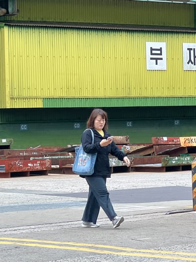 In Geoje-si, South Korea, a woman walks on a paved street, holding a smartphone. She is wearing a dark jacket, dark pants, and grey sneakers. A blue denim bag with red and white lettering is slung over her left shoulder. Behind her, a corrugated metal structure painted yellow and green features Korean characters and lettered signs. In the foreground and midground, stacks of industrial metal materials are visible. The scene appears to be an outdoor industrial or commercial area.