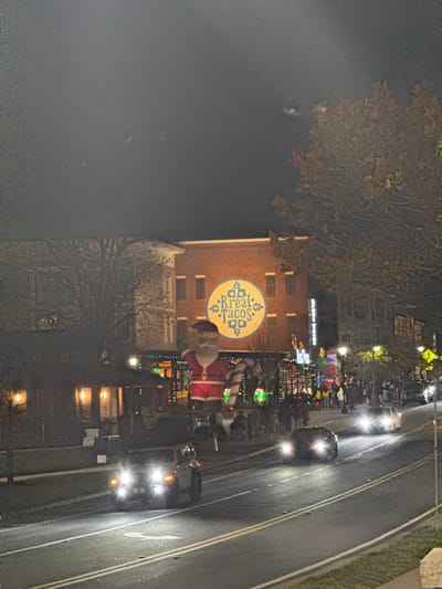 A festive night scene unfolds with "Kreal Tacos" displayed prominently on a brick building. An inflatable Santa and candy cane stand alongside colorful holiday lights lining the street. Cars with headlights on drive along the adjacent road. Silhouetted figures gather on the sidewalk, suggesting a lively event. Trees flank the scene, adding depth to the nighttime setting.