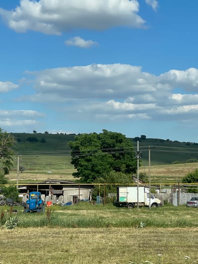 The image depicts a rural scene in Donskoe, Russia, under a bright, partly cloudy sky. The perspective is horizontal, showing a landscape with rolling green hills in the distance, fields of dry grass in the foreground, and a cluster of buildings and vehicles to the right.
Several objects are visible in the scene. In the foreground, a blue tractor is parked near a dilapidated barn-like structure with a corrugated metal roof. The structure appears to be used for storage, with various items, including what looks like blue plastic bins and other debris, piled around it. A white truck with a boxy cargo area is also parked near the buildings. In the middle ground, a line of utility poles with wires runs across the frame, dividing the sky from the landscape. The fields are a mix of green and dry, yellowed grass, suggesting it may be late summer or early autumn.
The sky is a vibrant blue with large, fluffy white clouds scattered across it, indicating fair weather. The lighting suggests it is daytime, likely mid-morning or afternoon. There are no people visible in the frame, and the overall mood of the image is one of quiet, rural existence. A sign with Cyrillic text is partially visible on the blue tractor, but the text is not legible. The scene evokes a sense of a functional, but perhaps slightly weathered, rural homestead.