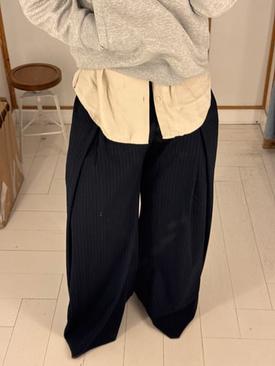 One individual, observed from the chest downwards, stands facing the camera in an indoor setting within the city of Gondomar, Portugal. The person is dressed in a grey long-sleeved sweatshirt layered over a light beige untucked button-up shirt; a single dark button is visible on the shirt. They are wearing dark navy or black wide-leg trousers with fine vertical pinstripes. The floor is composed of light-colored, rectangular planks, indicative of wood or laminate. To the left of the individual, a white metal stool with a round wooden seat is partially visible, positioned in front of a brown cardboard box. Further left, a light-colored wooden frame is present. To the right, a reflective surface, consistent with a mirror, features a thin light-colored frame. A fragment of blue denim material is also visible in the lower right background.