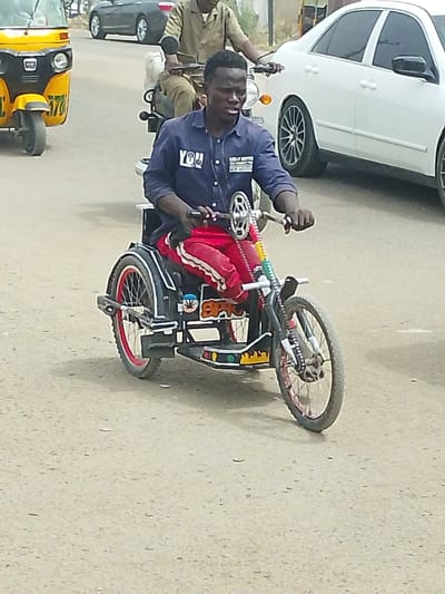 This image captures a street scene in Daurawa, Nigeria. The central figure is a young adult male, dark-complexioned with short dark hair, operating a custom-built, hand-powered tricycle. He is wearing a dark blue long-sleeved collared shirt with "YOU" printed in white on the left chest and additional white text ("KEEP IT REAL," "NEW MONEY") on the right, paired with red pants featuring light-colored side stripes. His hands grip the cranks, propelling the vehicle forward.
The tricycle is predominantly black with white accents and red-rimmed rear wheels. It features a visible chain drive system and a decorated side panel displaying "SPARO" in orange/yellow letters and a green cannabis leaf graphic. The front fork area also shows colorful decorative elements.
In the immediate background, a male individual in a light brown uniform-like outfit is seen operating a motorcycle, following behind the central figure. To the left, a yellow motorized tricycle (keke napep) is partially visible, oriented in the same general direction. To the right, a white sedan with dark-tinted windows and multi-spoke dark alloy wheels is present. Further in the background, another dark-colored sedan and indistinct vehicles are visible on the road.
The scene unfolds on a paved, light-colored road surface, possibly asphalt, under bright daylight conditions. All vehicles appear to be in motion or positioned within a traffic flow.