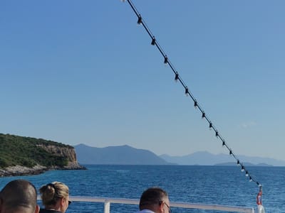 The image depicts a view from a boat on a clear day with blue skies and calm blue water. In the foreground, the backs of three people's heads are visible, two in focus and one slightly out of focus on the left. The person in the middle has blonde hair styled in a bun. A string of lights hangs diagonally across the upper right portion of the frame. In the background, a green, rocky coastline with a cliff face dominates the left side, with mountains visible in the distance across the water. The scene appears to be the coast of Pelasgia, Greece, possibly near a port or scenic area.