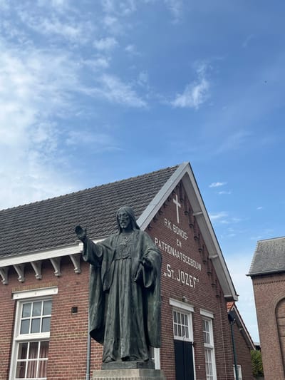 In Baarle-Nassau, Netherlands, a bronze-colored statue of Jesus Christ stands centrally on a light-grey concrete pedestal. The statue depicts Christ in a long, flowing robe, with a prominent Sacred Heart emblem on his chest. His right arm is raised with an open palm in a gesture of blessing, while the left arm is positioned slightly lower, holding a draped garment or scroll.
Behind the statue is a two-story, red brick building with a steep, dark grey tiled gable roof and white trim. White lettering on the upper facade of this building reads "R.K. BONDS- EN PATRONAATSGEBOUW "ST. JOZEF"" (Roman Catholic Association and Patronage Building "St. Joseph"), surmounted by a white cross. The building features multiple rectangular windows with white frames and dark lower panels on its ground floor. To the right of the main building, a section of another red brick structure with a dark roof is partially visible. The scene is set under a clear blue sky dotted with scattered white clouds.
