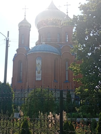 The image captures a view of a large, brick Orthodox church with golden domes, characteristic of religious architecture in Pokrov, Ukraine. The church features multiple domes of varying sizes, topped with crosses, and accented by blue roofing. A mosaic of Jesus Christ is visible on the side of the main structure, with accompanying text in Cyrillic script below it. The building is partially obscured by lush green trees and a black wrought-iron fence in the foreground, which encloses a garden area with various plants and flowers. The time of day appears to be daytime, with bright sunlight casting a haze over the scene, suggesting it could be a warm, sunny day. The overall impression is one of tranquility and solemnity, with the vibrant greenery and imposing religious structure creating a picturesque setting. No specific activity or event is depicted, focusing instead on the architectural grandeur and the surrounding natural elements.