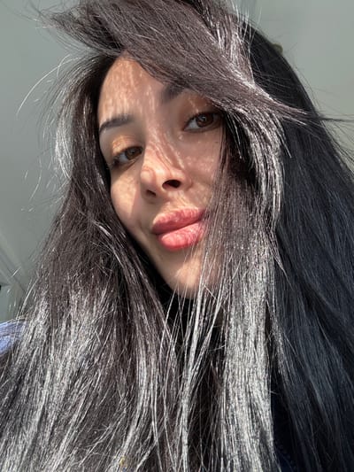 A close-up, upward-angled portrait shot depicts a female subject with long, dark hair, dark-brown eyes, and full, pink-tinted lips forming a slight pout. She is looking directly at the camera. Strong, directional sunlight from the upper left casts distinct, patterned shadows, resembling a lattice or blind, across the left side of her forehead, cheek, and nose. The sunlight also creates prominent specular highlights on her dark hair, making sections appear shiny and silvered. A dark collar is visible at her neck, and a portion of a blue garment is seen on her left shoulder. The background is a brightly lit, out-of-focus, light-colored interior wall. The scene is located in the city of Molodezhnyi, Russia.