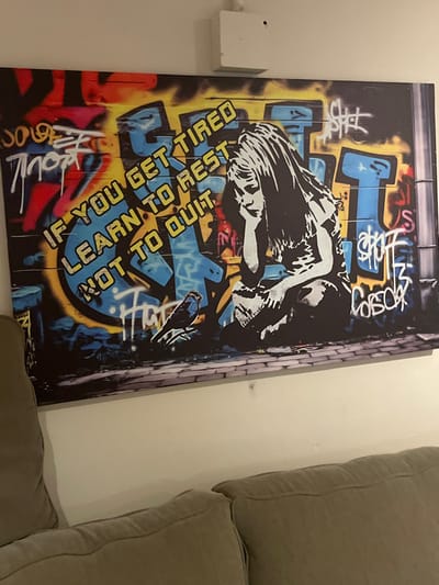 A graffiti-style print leans against a beige wall, partially obscured by a green cushioned couch. The print features a monochrome figure against a backdrop of blue, yellow, and black graffiti. Text in bold blue and yellow reads, "IF YOU GET TIRED LEARN TO REST NOT TO QUIT". A white electrical box is mounted on the wall beside the print. A brick effect design edges the print.