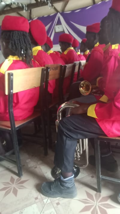 A group of people wearing red jackets and red berets sit on wooden chairs. A man holds a tuba. Most of the people have dark skin. The group appears to be in a band or a musical ensemble. The scene takes place indoors, with a tiled floor and a wall with purple decorations in the background.