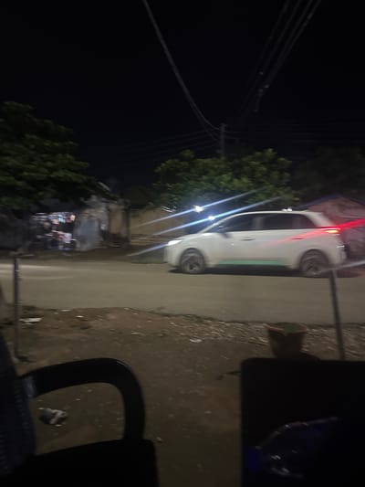 The image depicts a dimly lit street scene at night in Abuja, Nigeria. A white car is captured in motion, its headlights and taillights creating streaks of light across the asphalt. The car appears to be a hatchback with a distinctive green accent strip along its lower body. In the background, trees with dense foliage line the street, and structures that resemble buildings or stalls with illuminated signs are visible. The foreground shows a portion of a dark chair and a table, with a plastic bottle on the table. The ground is a mix of dirt and paved surfaces, with debris scattered around. The overall impression is that of a busy, albeit somewhat unkempt, urban environment at night.