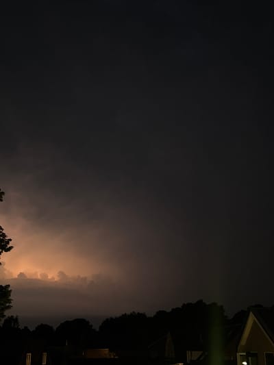 A photograph taken at night in the city of Putten, Netherlands, captures a dramatic sky illuminated by lightning. The horizon is dark with silhouetted trees and the rooftops of buildings. In the distance, a cluster of clouds is lit from within by a bright, orange glow, indicating a lightning strike. The foreground is dominated by a dark sky, with the lower portion of the image revealing the dark outlines of trees and houses, with some windows lit from within.