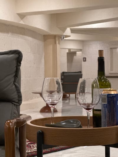 This indoor shot shows a wine cart with a wine bottle, a can, and two wine glasses. The wine cork has the word "KOCABA" on it. The can says "Edelmeister." The setting is a room in what appears to be a hotel or apartment in Ürgüp, Turkey, given the characteristic stone walls and archways. The room is furnished with a gray chair or couch. The scene is lit by natural light.