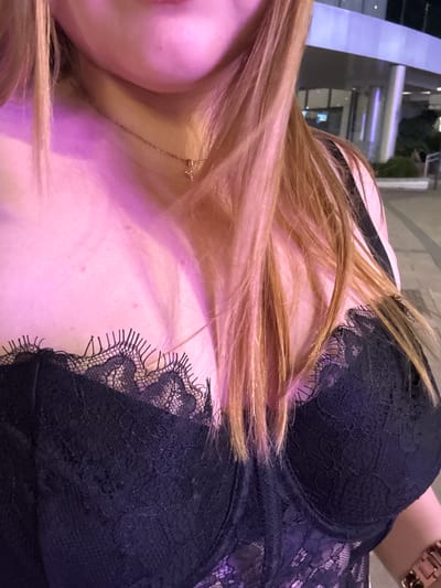 The image shows a close-up, top-down view of a person's chest and neck, with their hair cascading over their shoulder. They are wearing a black lace bra. A delicate gold necklace with a small charm is visible around their neck. The background is blurred, but appears to be an indoor setting with some ambient lighting, possibly with purple and pink hues reflecting on the skin. The person's skin has some freckles and small blemishes. The overall impression is intimate and personal. There is no visible text in the image. The location context provided (Alcorcón, Spain) does not offer any specific visual cues within the image itself.