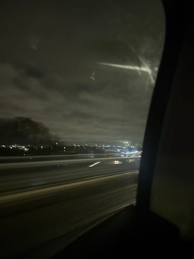 A nighttime view from inside a vehicle, likely a car, shows a rain-streaked windshield. Streetlights and the glow of traffic are visible in the distance, distorted by the water. To the right, the blur of passing highway lanes. The interior of the car, specifically the roof and side, frames the scene. The overall impression is one of driving in heavy rain at night.