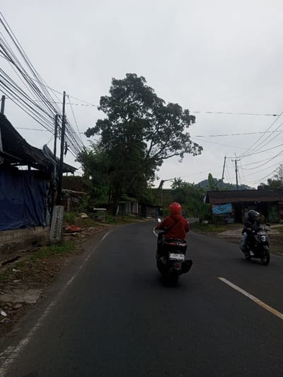 The image depicts a street scene in the city of Sat Thay (East), Myanmar, under an overcast sky. Two individuals are operating motorcycles on a dark asphalt road that curves slightly to the right, moving away from the viewer. The primary motorcyclist in the foreground wears a red helmet and a red hooded jacket, riding a dark scooter with the visible license plate F 4507 MBG. A second motorcyclist, wearing a dark helmet and dark clothing, is visible further ahead on the right side of the road, riding a dark motorcycle.
The road features a solid white line marking the right shoulder and a broken white center line further in the distance. On the left side of the road, multiple black power lines are densely strung between several utility poles. Behind these poles, a structure is partially covered by a dark blue tarp, adjacent to a weathered white sign post. Beyond this, an earthen mound with sparse vegetation rises, and small, simple structures with corrugated roofs are visible. A large, prominent tree with dense green foliage stands behind the structures on the left, near the center of the image.
On the right side of the road, a building with a dark corrugated roof displays a light blue tarp or banner on its front facade. Further in the background on the right, additional structures and green hills are visible. The overall environment suggests a semi-rural or village setting with typical local infrastructure.