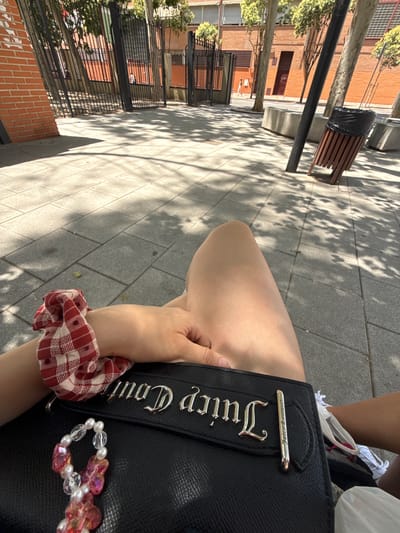 The image shows a person's legs and hands resting on a black handbag. The handbag has silver "Juicy Couture" branding. A red and white checkered scrunchie is on the person's wrist. The person is seated on a paved area in an outdoor urban setting. In the background, there are brick buildings, a metal fence, trees, and a trash can. The sun is casting dappled shadows on the ground, suggesting it is a sunny day. The presence of brick buildings and trees indicates an urban environment. Specifically, this scene is likely located in Alcalá de Henares, Spain, given the architectural style and vegetation.