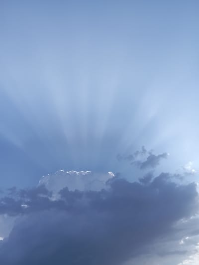 The image shows a vast, pale blue sky with crepuscular rays emanating from behind a large, cumulonimbus cloud formation. The cloud is dark and appears heavy, with bright white highlights along its upper edges where sunlight breaks through. Smaller, wispy clouds are scattered across the sky. The scene suggests a late afternoon or early evening with dramatic lighting conditions. The geographical location of Beruniy, Uzbekistan, is not evident from the image itself.