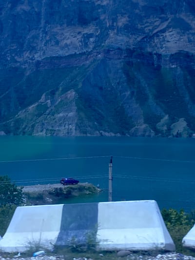The image captures a panoramic view of a mountainous landscape with a body of water. In the foreground, a white and gray concrete barrier is visible, partially obscured by dry grass and debris. Beyond the barrier, a turquoise body of water stretches across the frame, reflecting the dark blue hues of the towering mountains that dominate the background. The mountains are rugged and appear to be covered in sparse vegetation on their lower slopes, transitioning to barren rock faces higher up.
On a small, rocky outcrop situated near the water's edge, a dark purple car is parked. The car appears to be a sedan with a spoiler on the trunk. A single utility pole stands nearby, with electrical wires stretching horizontally across the scene. A person, appearing as a small, silhouetted figure, is visible near the water's edge by the car. The overall lighting suggests it is either early morning or late evening, contributing to the dark, moody atmosphere of the scene. The location is identified as Irganai, Russia.