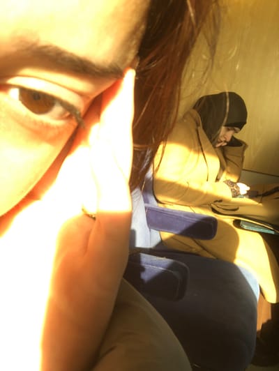 The image depicts an interior scene within a public transport vehicle, such as a train or bus, observed in the city of Dettwiller, France. In the foreground, heavily illuminated by direct sunlight, the right eye, eyebrow, cheek, and a section of brown hair of one individual are visible. A hand is positioned near the subject's temple, contributing to the bright overexposure. In the background, a second individual is seated in a blue upholstered chair. This person wears a black head covering and a light brown/tan winter coat. They are looking downwards at an object in their lap, with their left hand, adorned with a dark, possibly lace-trimmed glove, visible near the object. Strong natural light creates significant highlights and shadows throughout the scene, particularly on the seated individual and the vehicle's interior.