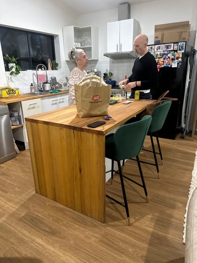 In Bonville, Australia, a couple stands in a kitchen. The woman, with grey hair styled in a bun and wearing a patterned dress, stands near a sink and a window. The man, bald and wearing a black sweater, is pouring a drink from a can into a glass. A wooden kitchen island with three green bar stools occupies the foreground, with a grocery bag from IGA placed on it. The kitchen is equipped with white cabinets and appliances, and the floor is made of wood-look laminate. The overall scene depicts a casual domestic moment.