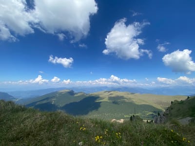 A scenic, wide-angle landscape photograph captures a lush mountain vista under a bright blue sky dotted with fluffy white clouds. The foreground is dominated by tall, green grass, interspersed with small yellow wildflowers, giving a sense of wild, untamed nature.
Beyond the grassy slope, rolling green mountains stretch into the distance. The mid-ground features a prominent mountain ridge with a road winding along its crest. Deep shadows are cast across the slopes, indicating the sun is positioned to one side, likely late morning or early afternoon. Patches of dark green forest are visible on the mountainsides, contrasting with the lighter green of the grassy areas.
In the background, a series of layered mountain ranges fade into a hazy blue, suggesting a vast and expansive landscape. The sky is a vibrant cerulean, with large, billowy cumulus clouds casting shadows on the mountains below. The overall impression is one of a serene, sunny day in a mountainous region. There are no people or man-made objects clearly visible, aside from the faint road. The location is suggested to be Santa Cristina Gherdëina, Italy, a region known for its stunning Dolomite scenery. The mood is peaceful and awe-inspiring, showcasing the natural beauty of the mountains. There is no visible text in the image.