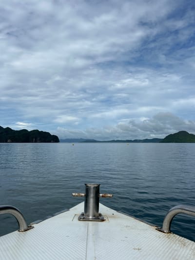 A boat is moving through calm, dark blue water towards a chain of verdant, hilly islands under a cloudy sky. The foreground of the image shows the bow of the boat, a white surface with a textured grip, and a shiny metal cleat. On the left and right sides of the bow, there are curved metal railings. In the distance, the islands are covered in lush green vegetation, and some have rocky cliff faces. The water is slightly rippled, and there are a few yellow buoys visible on the surface of the water between the boat and the islands. The sky is filled with a mix of white and grey clouds, with patches of blue sky visible. The scene depicts a journey through a natural, aquatic environment.