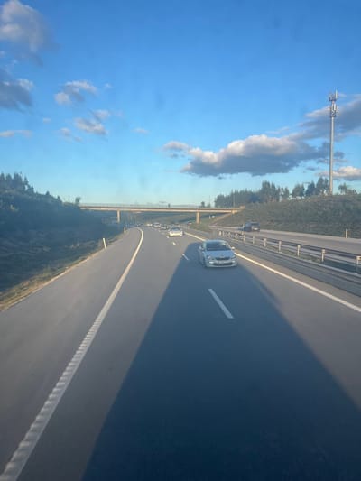 The image depicts a multi-lane highway in the region of Pombal, Portugal, under a clear blue sky with scattered white clouds, illuminated by low-angle sunlight, indicative of late afternoon or early morning. The perspective is from a moving vehicle. The highway features at least two lanes of travel in the foreground, separated by solid and dashed white lines, with a rumble strip bordering the left lane. A prominent, elongated dark shadow extends across the right lane from an unseen object to the right. A silver Peugeot sedan, license plate 71-WR-81, is positioned in the right lane in the mid-ground. Several other sedans, both light and dark-colored, are observed in the left lane further down the road. An overpass, constructed of concrete or stone with a metal barrier, spans the highway ahead. Both sides of the roadway are flanked by rising terrain densely covered with dark green vegetation. On the far right, beyond the tree line, a tall cellular communication tower equipped with multiple antenna arrays is visible against the sky. No individuals are discernible in the scene.