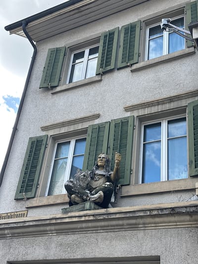 A statue of a Native American man, adorned with traditional clothing and a headdress, sits on a ledge outside a building in Wald (ZH), Switzerland. He is positioned to the left of a window, with his left arm resting on what appears to be a wolf or dog. His right arm is raised, with his thumb extended upwards in a gesture of approval. The statue is placed on top of a green crate. The building has a light-colored facade, and features several windows with green shutters, some of which are open, revealing the sky and clouds. A street sign that reads "Sösslalstrasse" is visible on the ledge below the statue. The sky is overcast with patches of blue sky.