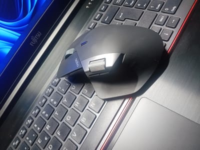 An overhead shot shows a black computer mouse resting on a laptop keyboard. The laptop has a partially visible screen on the left displaying a blue abstract pattern. The keyboard is dark gray with white lettering and a red trim. The mouse has a silver scroll wheel and additional buttons. The image is taken from an angle, highlighting the textures of the keyboard and the mouse. The scene is indoors, likely in an office or home environment, and does not contain any information about the city of Bohodukhiv, Ukraine.