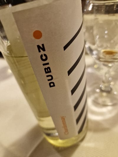 The image displays a close-up, slightly tilted shot of a clear glass bottle filled with a pale yellow liquid, identified as "Chardonnay" wine by its label. The bottle's white label features the prominent vertical text "DUBICZ" in black, topped by a small orange circular dot. Diagonal black stripes are visible on the right side of the label, and the word "Chardonnay" is printed in a smaller, orange-brown font below "DUBICZ." The bottle rests on a light-colored surface, presumably a tablecloth. In the out-of-focus background, a stemmed, empty or near-empty clear wine glass is partially visible to the right. The scene is illuminated by warm, artificial indoor lighting, creating reflections on the glass objects. This setting is located in the city of Odorheiu Secuiesc, Romania. No individuals are present, and no actions or interactions are depicted.