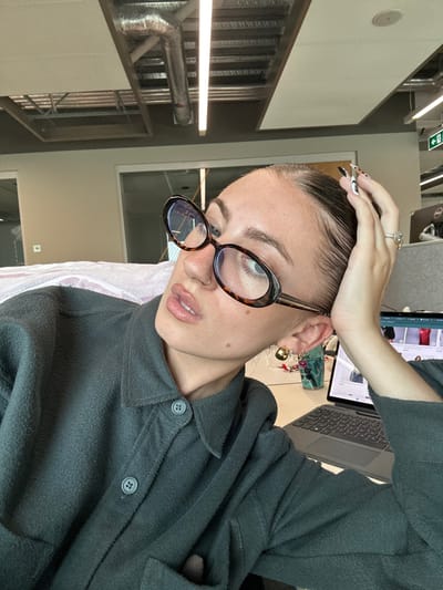 A female individual is depicted in an office environment in Garston, United Kingdom. She is seated, with her head tilted slightly upwards and to her right, gazing towards the upper left. Her dark hair is slicked back. She wears oval, tortoiseshell-patterned eyeglasses that show a blue light reflection, and a small hoop nose ring on her right nostril. Her lips appear glossy. She is dressed in a dark teal or muted green collared shirt with visible buttons. Her left hand, featuring long, patterned fingernails and a ring with a large central stone on the ring finger, rests on the top of her head. A distinct gold-toned earring, incorporating an additional colored element, is visible on her right ear.
On the white desk surface in front of her, a dark-colored laptop with a visible keyboard displays an e-commerce website on its screen. A clear or teal-colored beverage bottle, marked "DK KEEP," is positioned behind the laptop. The background reveals an open-plan office layout, characterized by exposed metallic HVAC ductwork, white rectangular ceiling panels, and linear fluorescent or LED lighting fixtures. Light-colored partition walls are visible, along with a partial view of a glass partition. Further in the background, a black Dell monitor and a green exit sign are discernible.